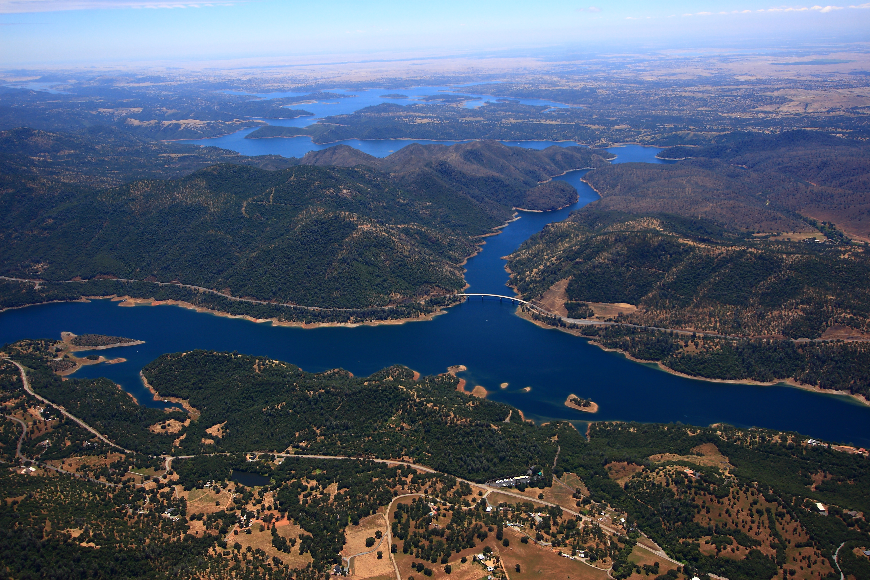 Aerial view of a long, winding lake with a bridge crossing it, surrounded by forested hills and scattered rural homes under a clear sky.